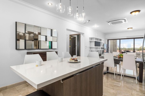 A modern kitchen with a white countertop, wooden cabinets, and a large mirror. There is a dining area with white chairs and a table, and a view of the city through large windows.