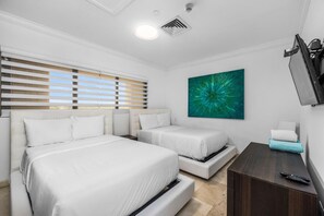Habitación de hotel con dos camas, un ventanal con persianas, un cuadro abstracto color turquesa, un televisor y un escritorio con un control remoto.