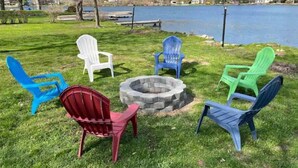 A lakeside seating area with Adirondack chairs in various colors arranged around a stone fire pit. - Beautiful 3 Bedroom Lakefront Home (Sweet Valley)
