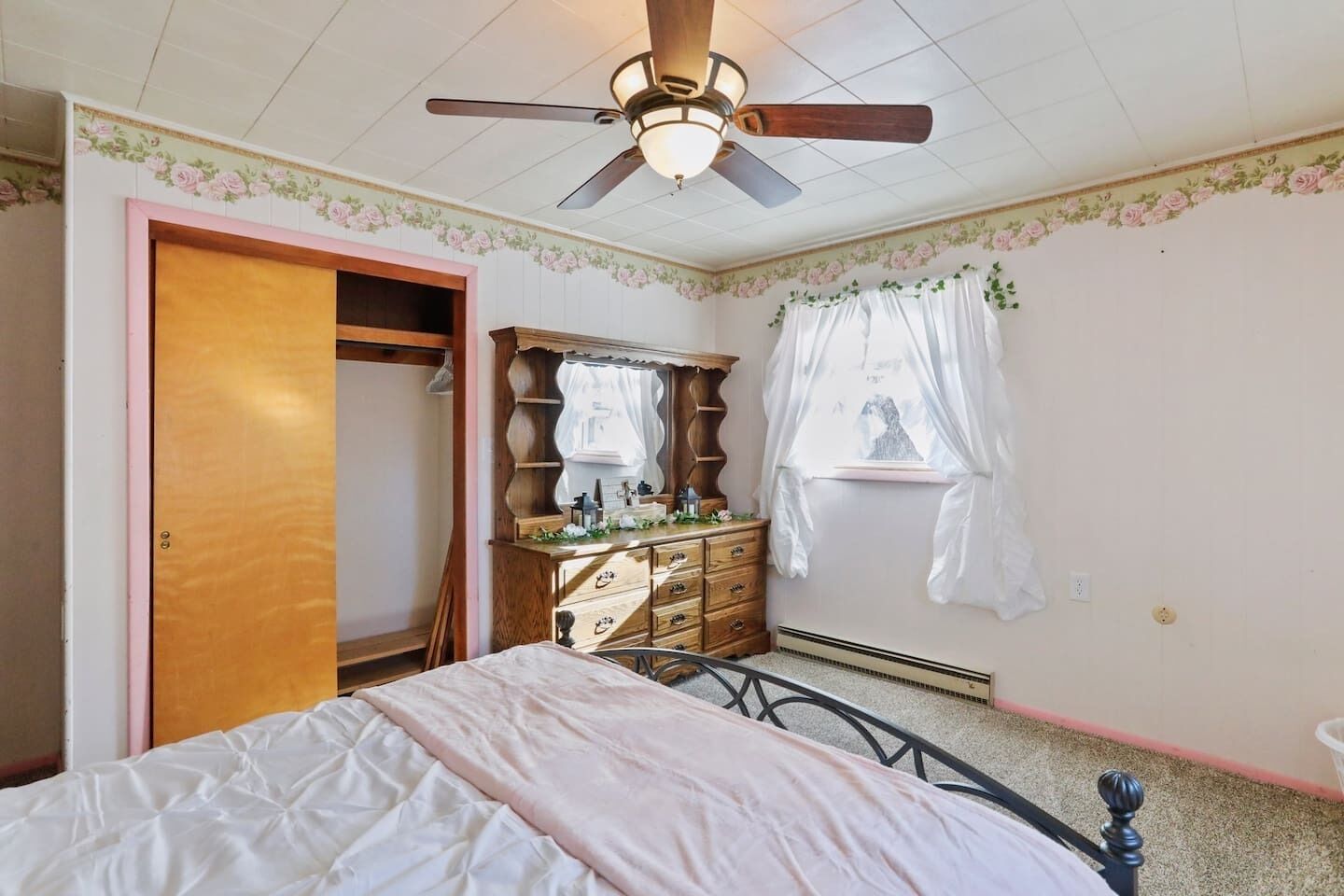 A bedroom with a bed, a dresser, a vanity, a ceiling fan, and a window with curtains.