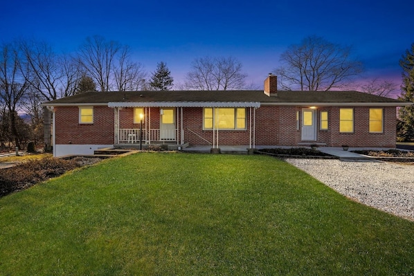 A two-story brick house with a front porch, a small front yard, and a gravel driveway.