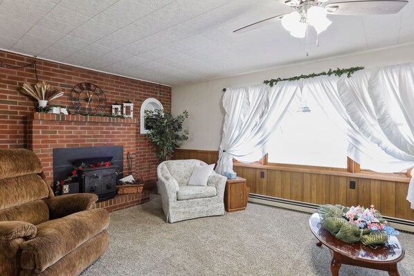 Un salon avec une cheminée en briques, un ventilateur de plafond, une fenêtre avec des rideaux blancs et une petite table agrémentée d’un arrangement floral.