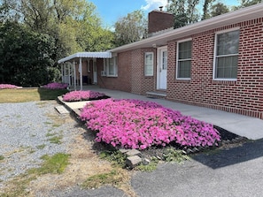 A brick building with a covered porch and pink flowers in the front yard.