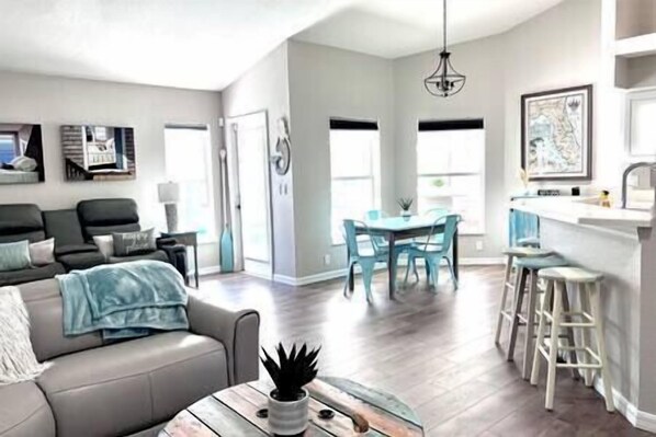 A spacious living area with a sofa, a dining table with chairs, a kitchen area with stools, and a large window providing natural light.