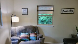 A living room with a grey armchair, a black sofa, a window with blinds, and two wall decorations. - Stock Cottage Cozy 2 BR, 2 Bath House next to Biology Trails and SAU (Ooltewah)