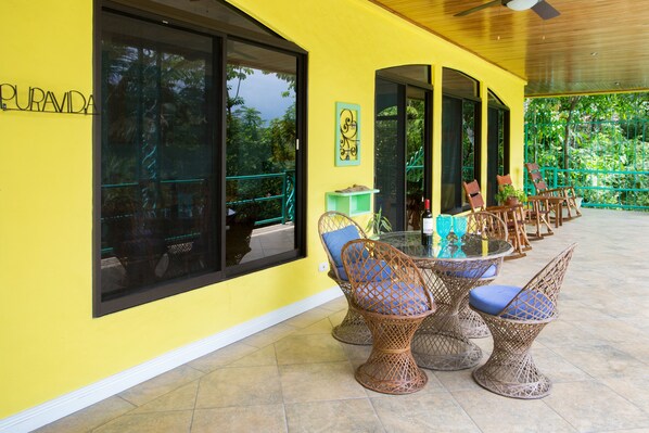 Un mur extérieur jaune avec de grandes fenêtres et une table en verre entourée de chaises en osier.