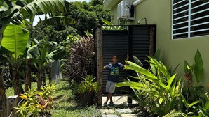 Un sendero lleva a una casa de color verde, rodeada de exuberante vegetación tropical y un cielo azul despejado.