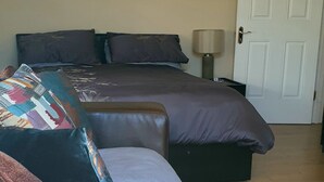 A room with a brown leather sofa, a bed with dark bedding, a lamp, and a door.