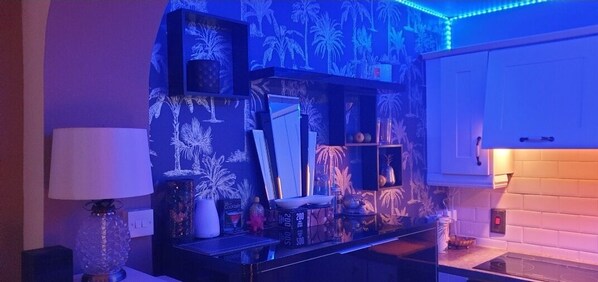 Habitación con zona de cocina que incluye un armario blanco, una encimera y un espejo decorativo. Se observan estantes con varios objetos y una pared con un diseño tropical.