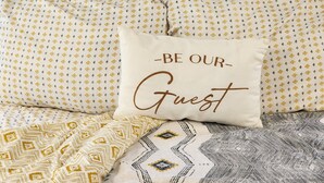 Una cama con ropa de cama estampada y una almohada con el texto "BE OUR GUEST".