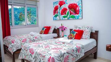 Habitación de hotel con dos camas, colchas estampadas con motivos florales, un cuadro de flores rojas y una ventana con cortina roja.