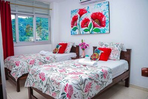 Habitación de hotel con dos camas, colchas estampadas con motivos florales, un cuadro de flores rojas y una ventana con cortina roja.