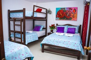 Una habitación con dos literas, cada una con una colcha azul estampada con flores y almohadas púrpuras. En la pared hay un cuadro de flores, un jarrón con rosas rojas y una cortina roja.