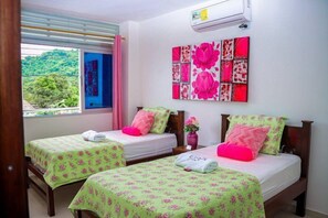Habitación de hotel con dos camas, cada una con una colcha estampada con motivos florales y almohadas rosadas. Se ve un ventanal con vistas a zonas verdes.