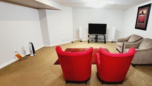Un salon avec un téléviseur à écran plat, deux fauteuils rouges, un canapé gris et un tapis rouge.