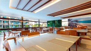 Un restaurante moderno con vista al océano, que cuenta con mesas y sillas de madera, ventanas amplias y un mural tropical.