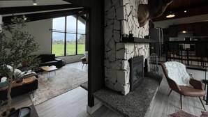 Un salon moderne avec une cheminée en pierre, une tête de cerf accrochée au mur et une grande fenêtre donnant sur un paysage verdoyant.