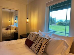 Ein Schlafzimmer mit einem großen Bett, einem Spiegel, Nachttischen und einem Fenster mit Blick auf Grünfläche.