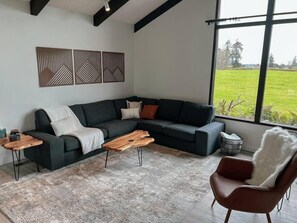 Ein modernes Wohnzimmer mit einem dunkelgrauen Ecksofa, einem Holztisch, einem roten Sessel und einem großen Fenster mit Blick auf ein grünes Feld.