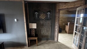 A room with a dartboard, a small table with a lamp, and a wooden floor. - Harbour Hutch  (St. Anthony)