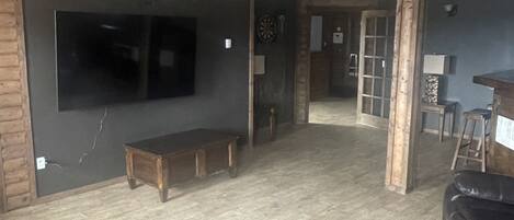 A spacious living room with a large flat-screen TV, a wooden bench, a black leather sofa, and a ceiling fan.