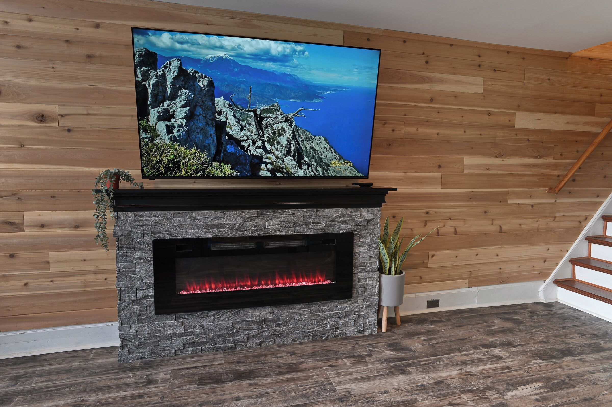 Uma sala de estar moderna com lareira de pedra, uma televisão de tela plana grande fixada em uma parede de madeira, e uma escada à direita.