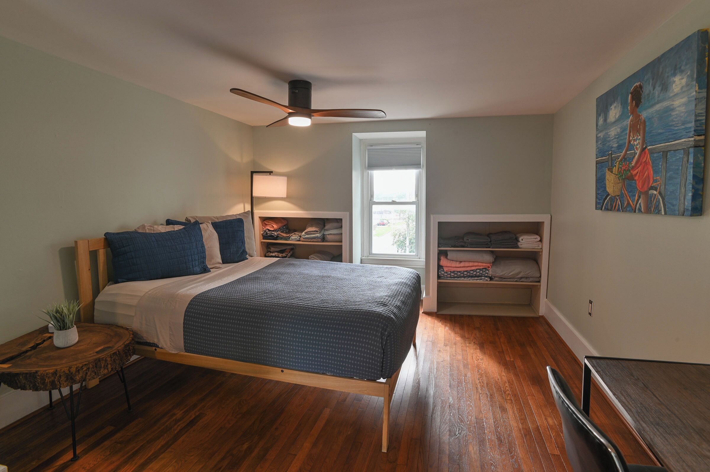 A bedroom with a bed, a ceiling fan, a window, a painting, and built-in shelving.