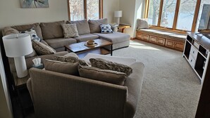 A living room with a sofa, armchairs, a coffee table, and a television. There is a window with a view of trees and a clock on the wall. - Private Lake Home, Game Room & Pontoon Rental on Keewahtin Lake (Forest Lake)