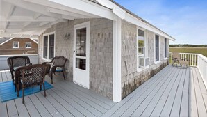 A covered deck with two wicker chairs and a table, a white exterior wall, and a view of a grassy field. - Oceanfront Family Retreat in Scituate with Private Beach, Expansive Porches, Outdoor Shower (Scituate)