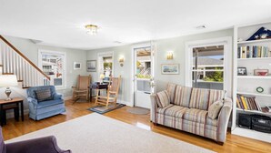 A spacious living room with a blue sofa, a striped sofa, and a wooden chair. There is a bookshelf filled with books and a small table with a lamp. A staircase is visible in the background. - Oceanfront Family Retreat in Scituate with Private Beach, Expansive Porches, Outdoor Shower (Scituate)