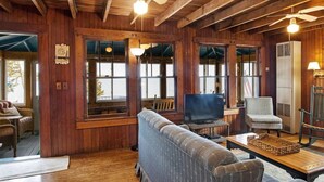 Ein gemütliches Blockhaus-Interieur mit Holzwänden, großen Fenstern, einem Fernseher, einer Couch und einem Tisch.