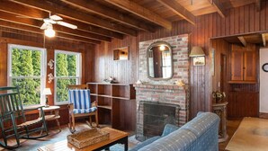 Ein gemütliches Wohnzimmer mit Holzwänden, einem Kamin aus Ziegeln, einem blauen Sofa, einem Schaukelstuhl und einem Esstisch.