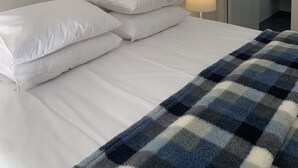 Siististi pedattu sänky, jossa on valkoiset lakanat ja ruudullinen peitto, yöpöytälamppu ja ovi, joka johtaa kylpyhuoneeseen.