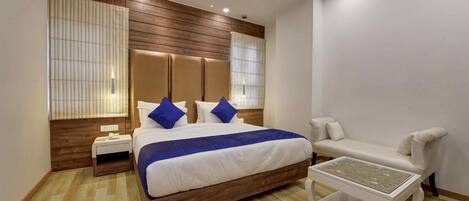 A modern hotel room with a wooden headboard, a large bed with blue pillows, a white sofa, and a glass coffee table.