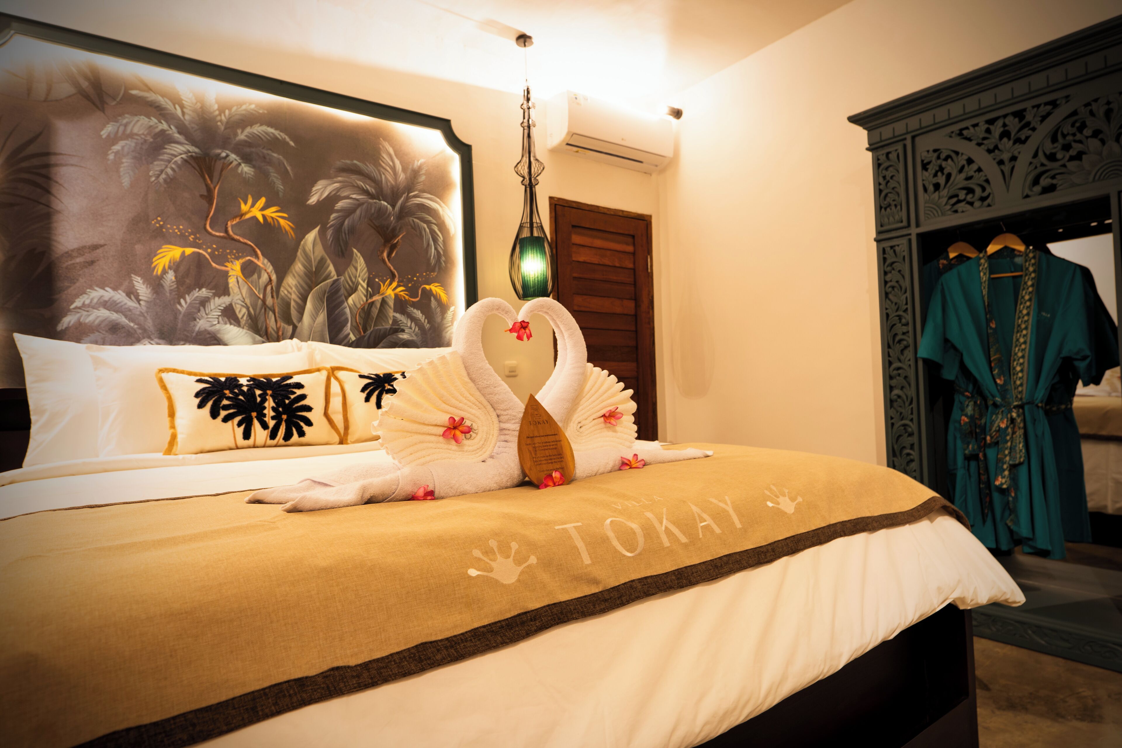 A neatly made bed with a towel swan, a tropical mural, and a teal robe hanging on a rack.