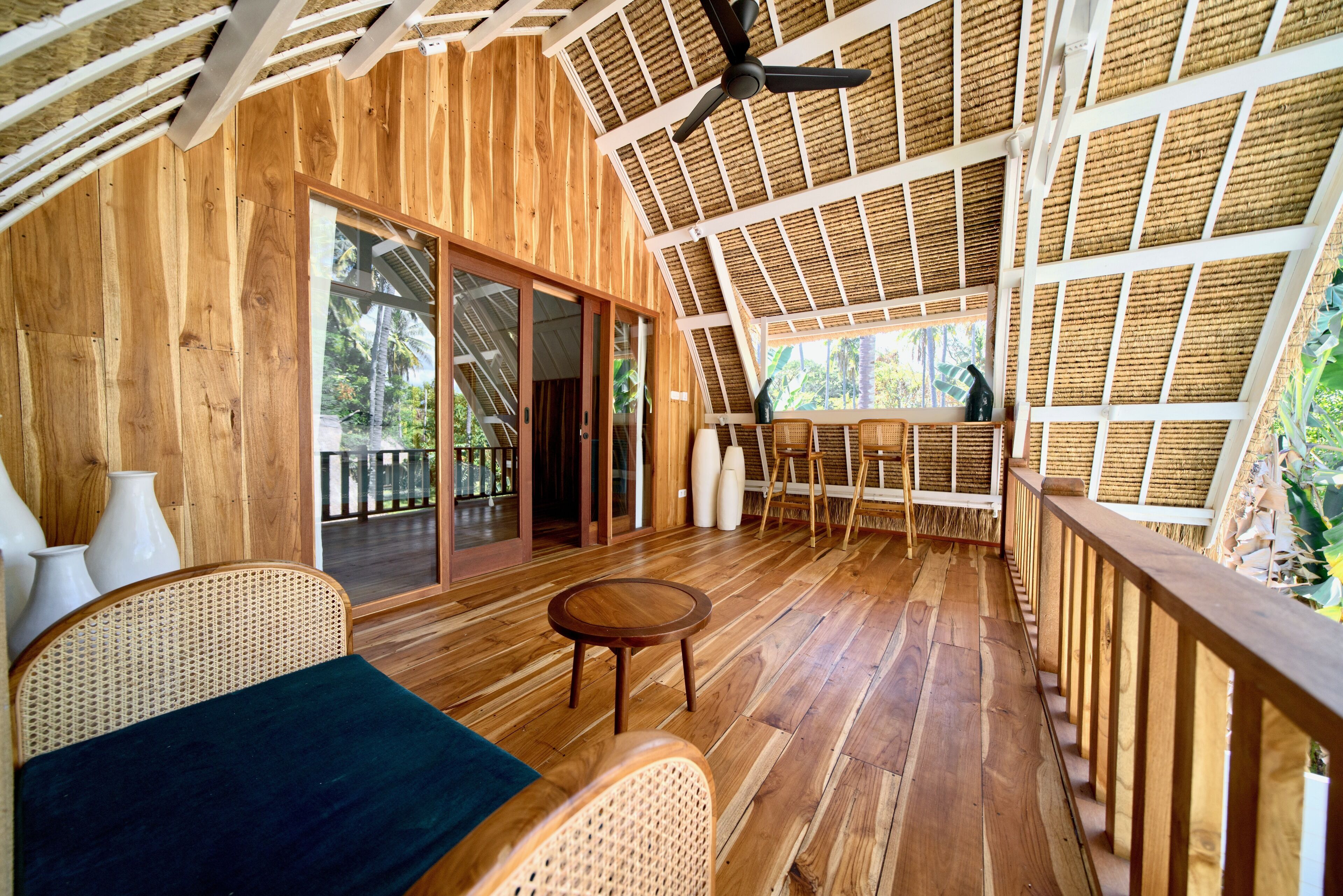 Un interior de madera con ventilador de techo, un amplio ventanal corredero, mobiliario de mimbre y vistas al exterior.