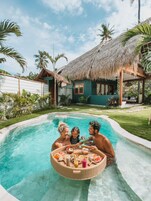 Un área de piscina con una estructura de techo de paja, rodeada de abundante vegetación y palmeras.