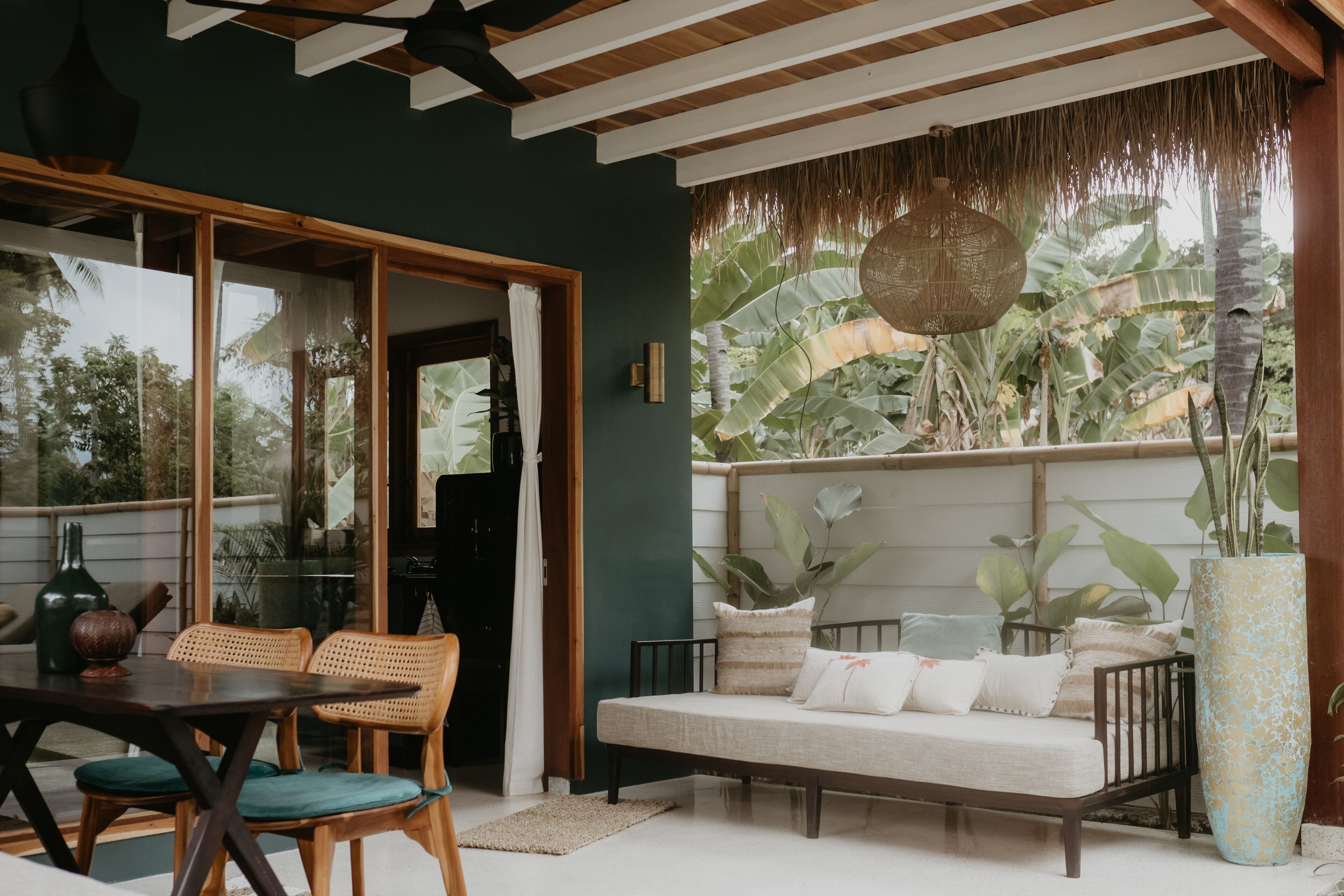 A room with a wooden ceiling, a sofa with cushions, a dining table with chairs, and a large plant in a vase.