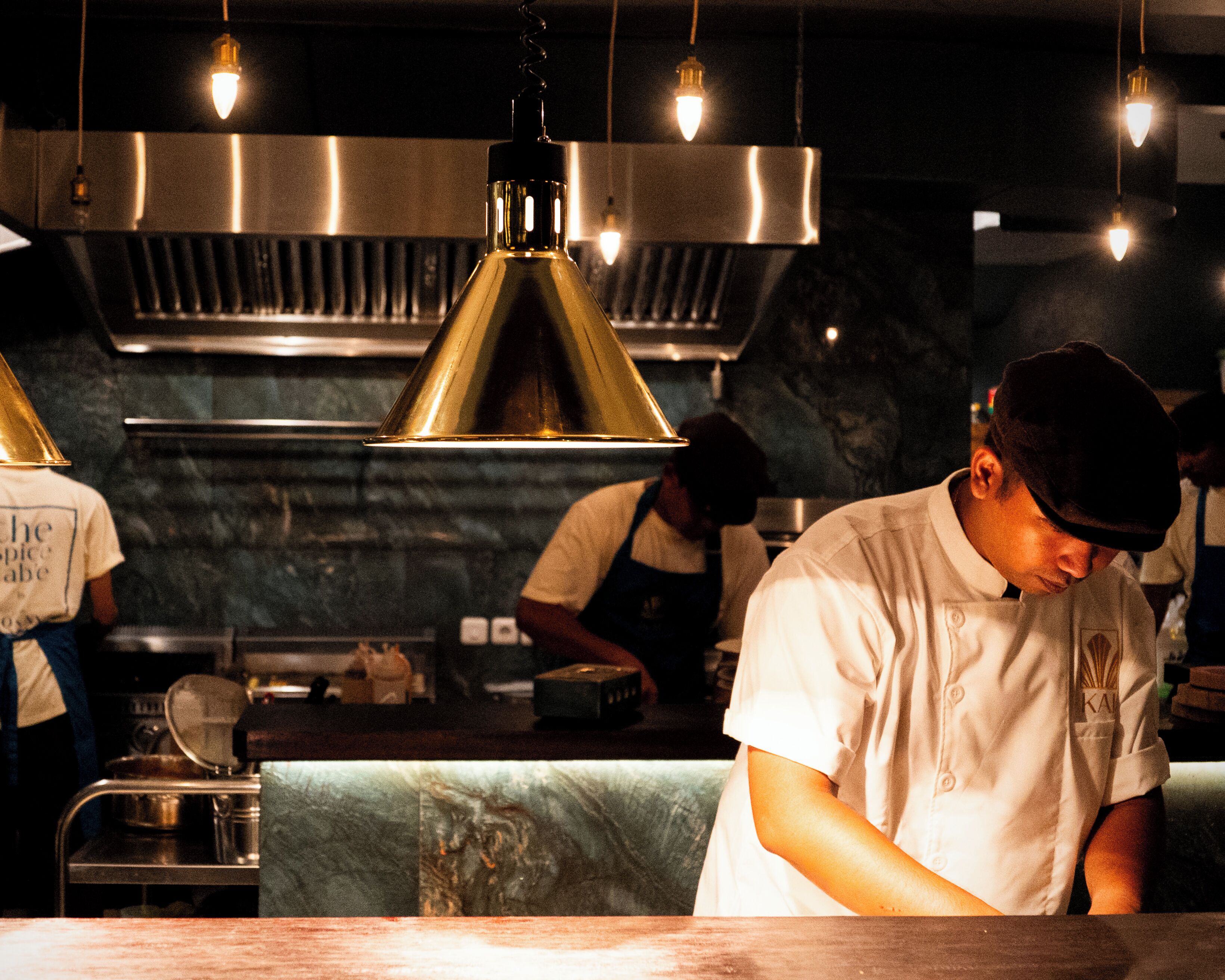 A chef in a professional kitchen with stainless steel appliances and pendant lighting.