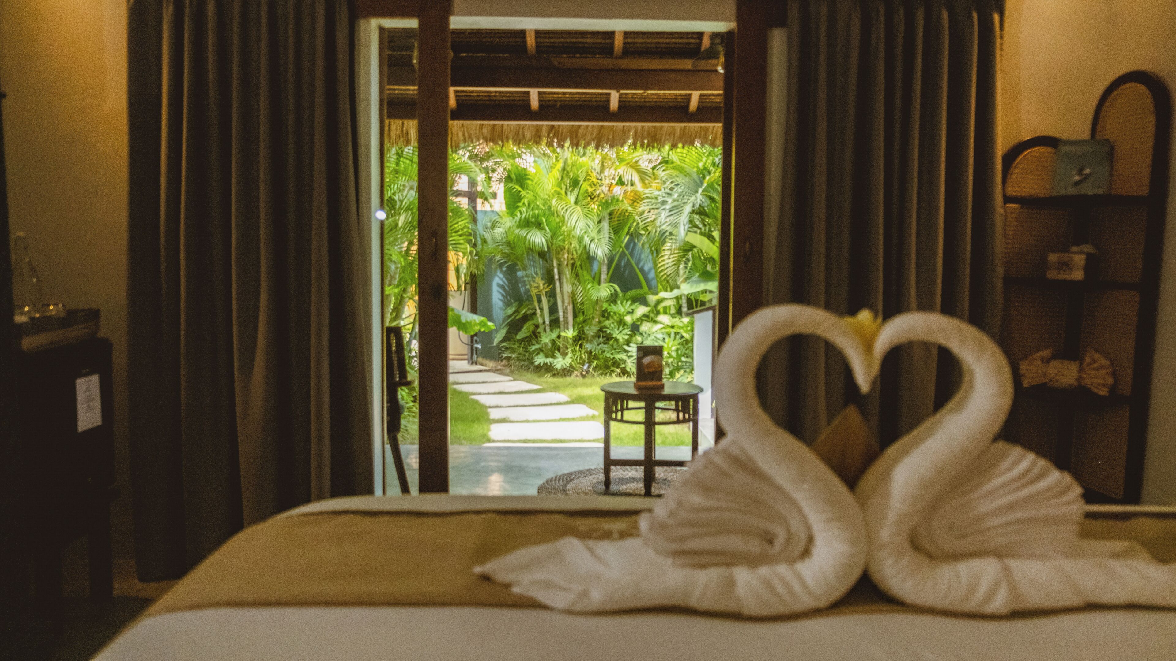 A bed with two swan-shaped towels, a small table, and a view of a tropical garden through a doorway.
