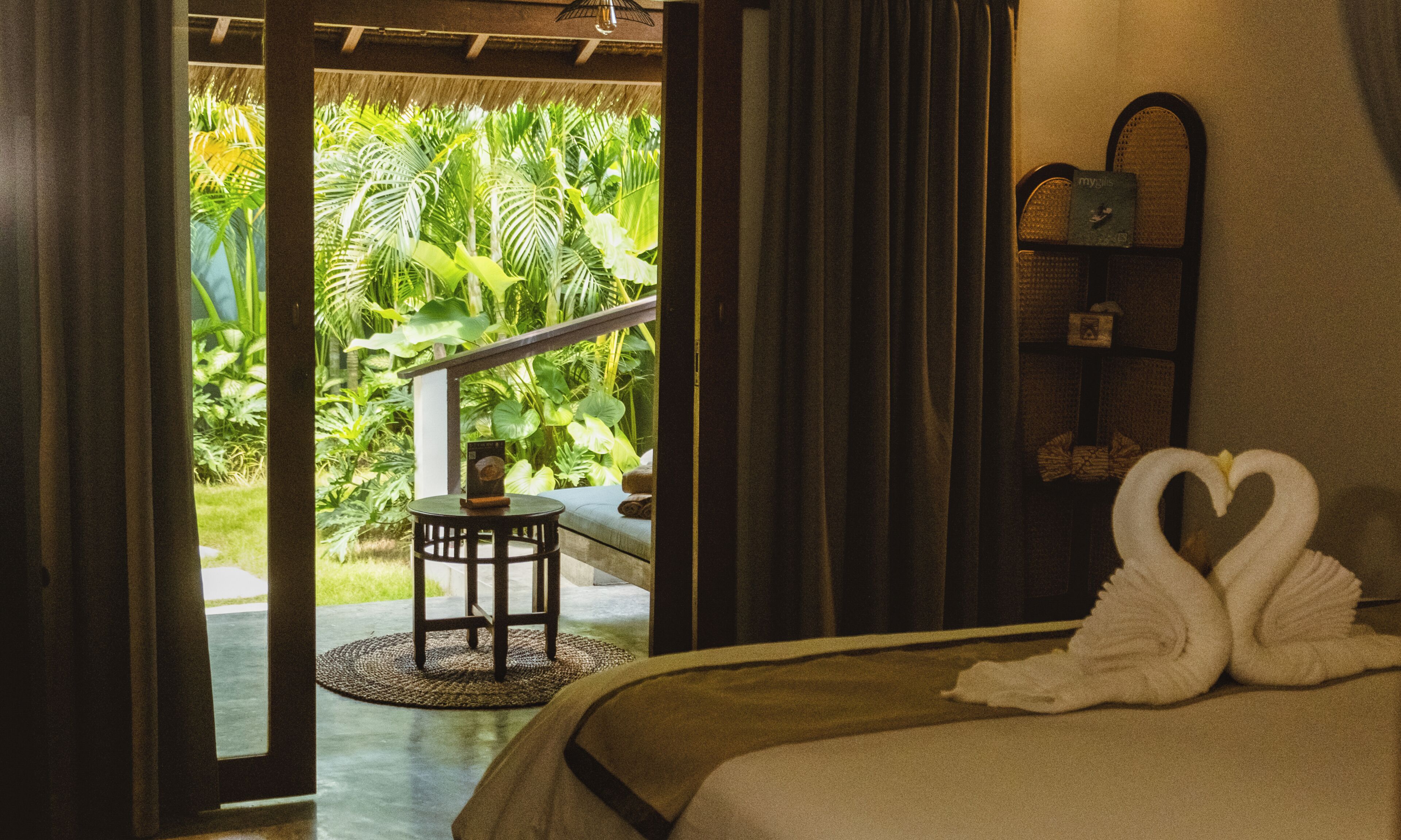 Habitación de hotel con cama, una mesita con silla, vista a vegetación exuberante a través de un ventanal y una toalla con forma de cisne sobre la cama.