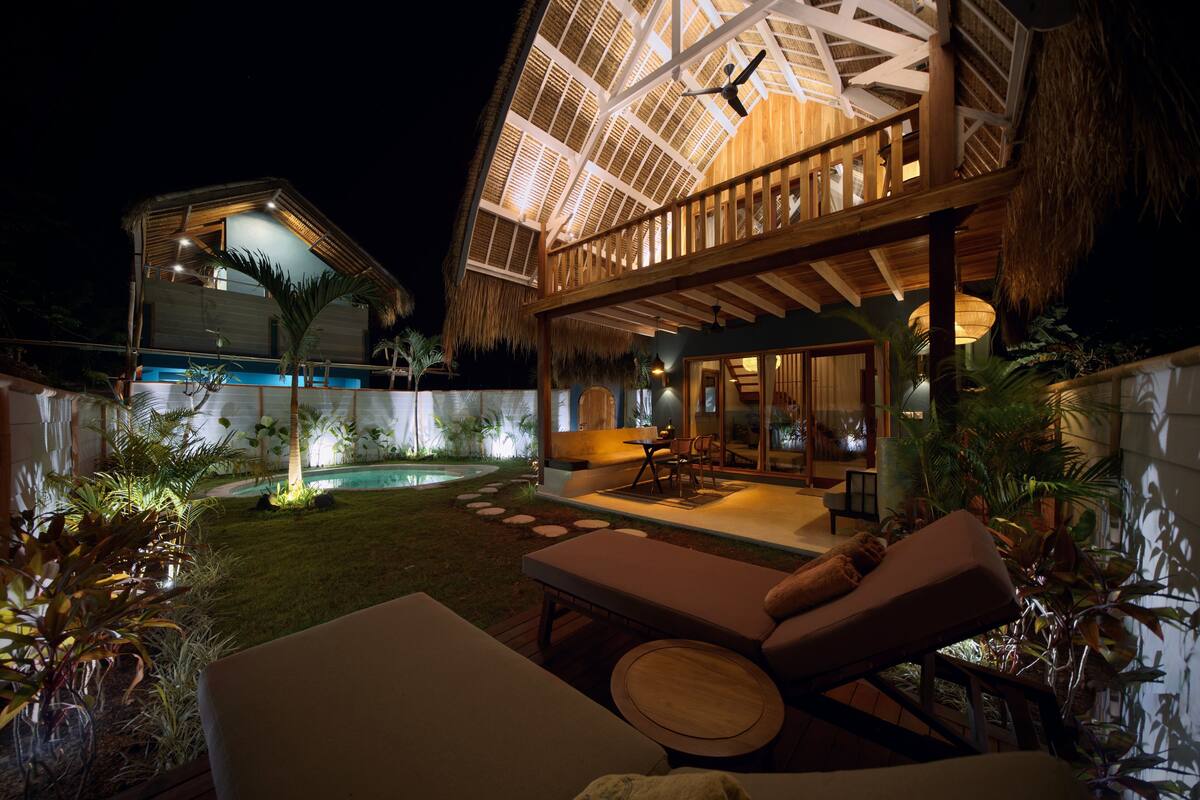 Un espacio exterior bien iluminado con piscina, sillas de descanso y una estructura de techo de paja.