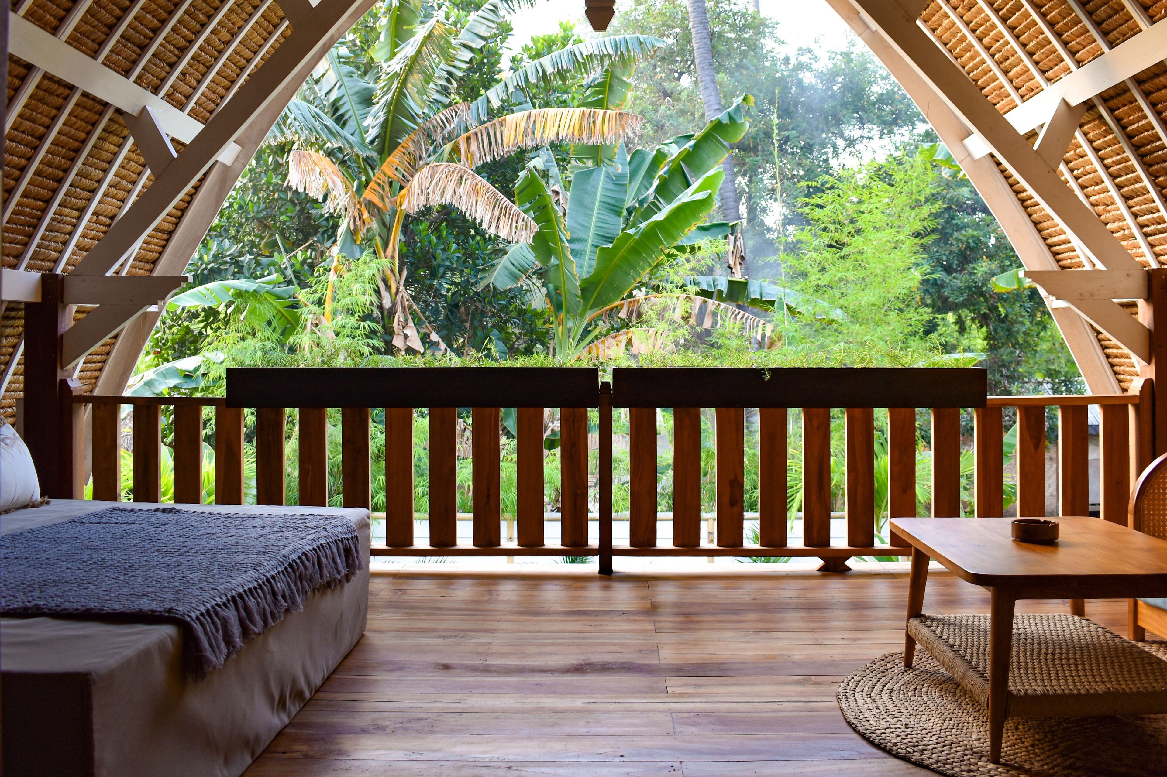 A wooden deck with a bed and a table, surrounded by lush greenery and a thatched roof structure.