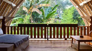 A wooden deck with a bed and a table, surrounded by lush greenery and a thatched roof structure.