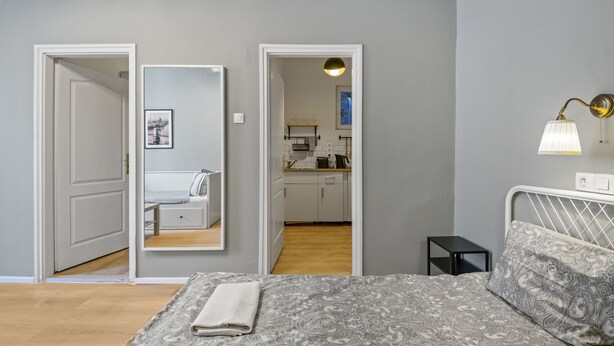 Quarto com cama, mesa de cabeceira, espelho e vista para uma cozinha e banheiro através de uma porta aberta.