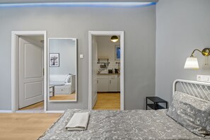 Quarto com cama, mesa de cabeceira, espelho e vista para uma cozinha e banheiro através de uma porta aberta.