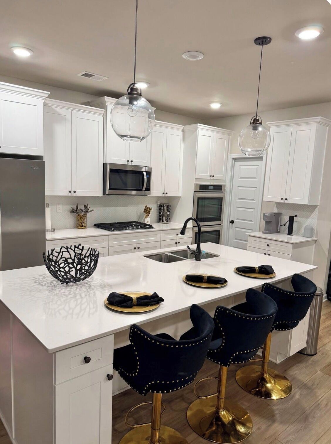 Una cocina moderna con muebles blancos, electrodomésticos de acero inoxidable, una isla central con taburetes y luminarias colgantes.