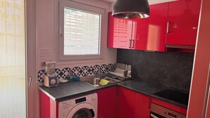 A compact kitchen with a washing machine, a sink, and red cabinets. - Exceptional Sea View – Terrace & Private Parking (ROSAS)
