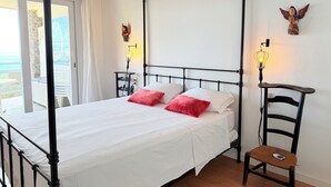 A four-poster bed with white linens, two red pillows, a nightstand with a lamp, and a chair with a remote. - Exceptional Sea View – Terrace & Private Parking (ROSAS)
