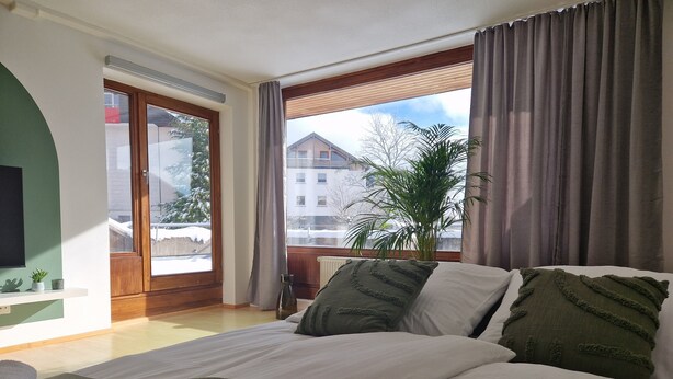 A hotel room with a bed, a potted plant, and a view of a building through the window.